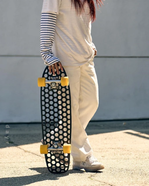 Woman in white outfit poses with a black perforated skateboard with yellow wheels on sunny concrete with a light gray wall backdrop.