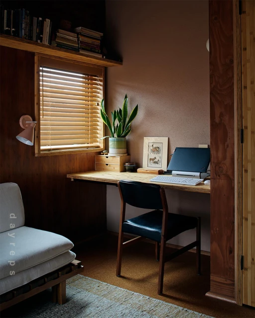 Indoor workspace featuring a wooden desk, chair, laptop, potted plant, and window with blinds creating a cozy and productive environment.