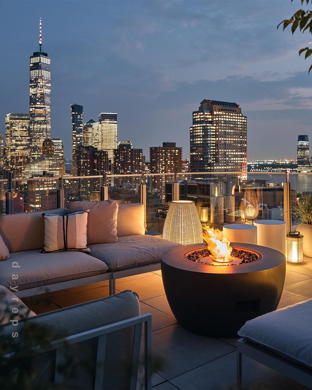 Rooftop patio with fire pit and lounge seating overlooking the New York City skyline at dusk, illuminated by building lights and the fire.