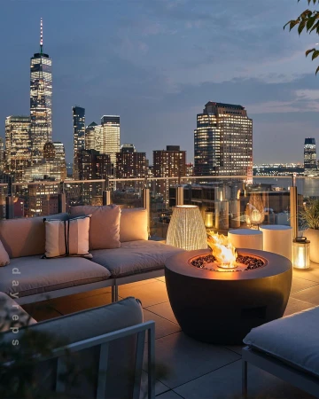 Rooftop patio with fire pit and lounge seating overlooking the New York City skyline at dusk, illuminated by building lights and the fire.