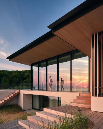 Exterior view of a modern home with a flat roof and floor-to-ceiling windows, featuring two people on the patio at sunset.