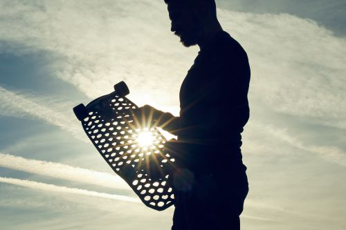 Silhouette of a man holding a skateboard against a bright sky with scattered clouds, backlit by the sun creating a lens flare.