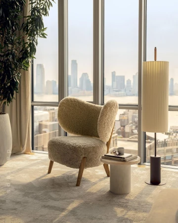Interior shot of a faux-fur chair next to a round side table and floor lamp in front of a large window with city skyline view.