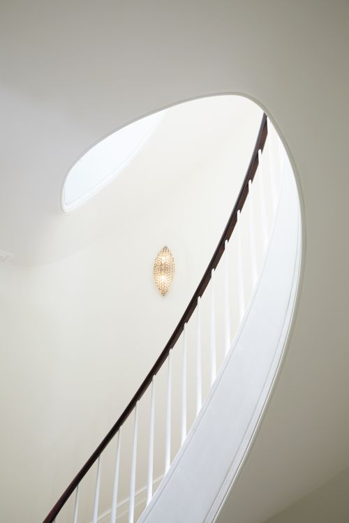 Curved white staircase with dark wood railing and white balusters, illuminated by a decorative wall sconce in a bright, modern home interior.
