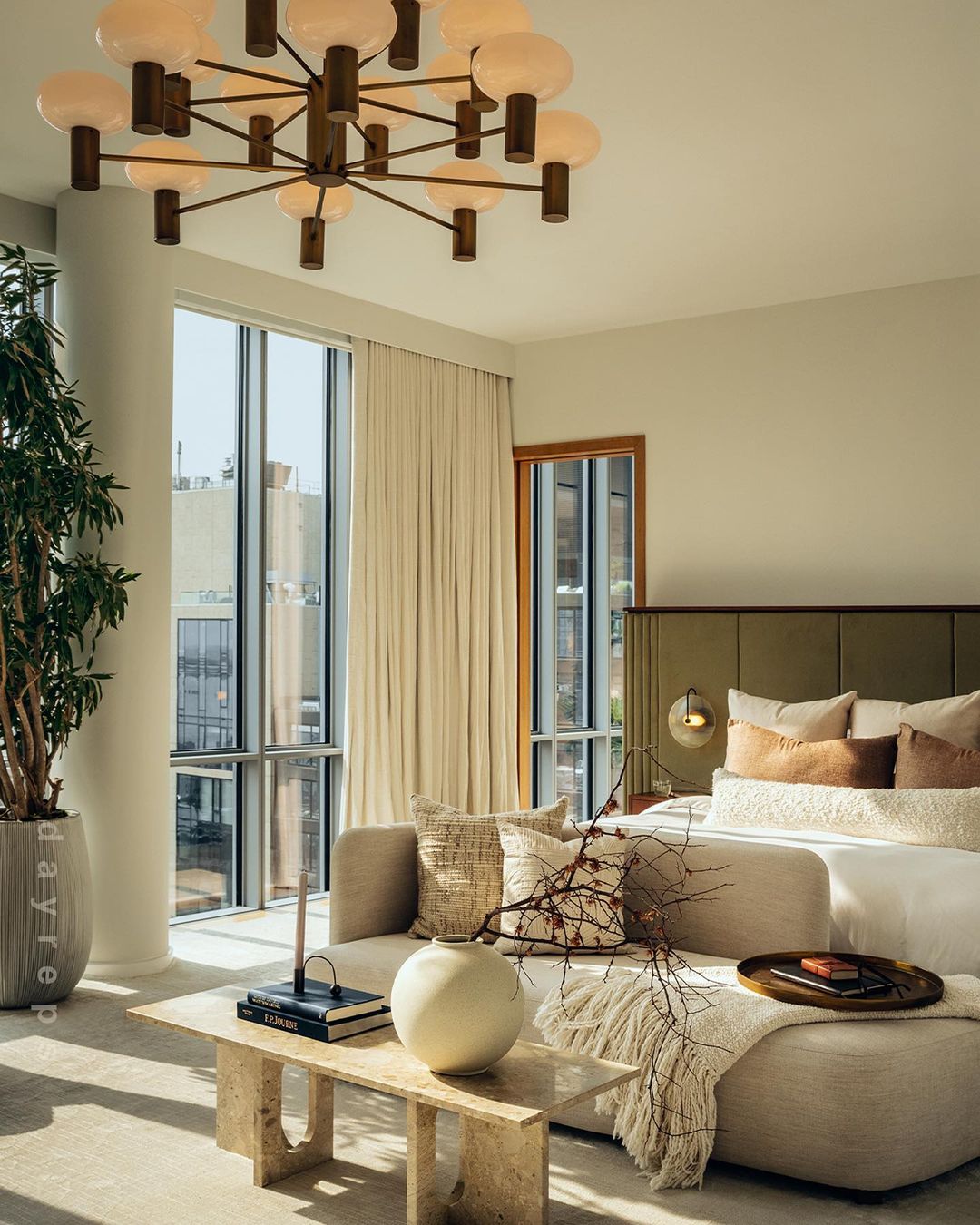 Well-lit bedroom with a large bed, a decorative couch, a table with books and a vase, and a potted plant near a bright window.