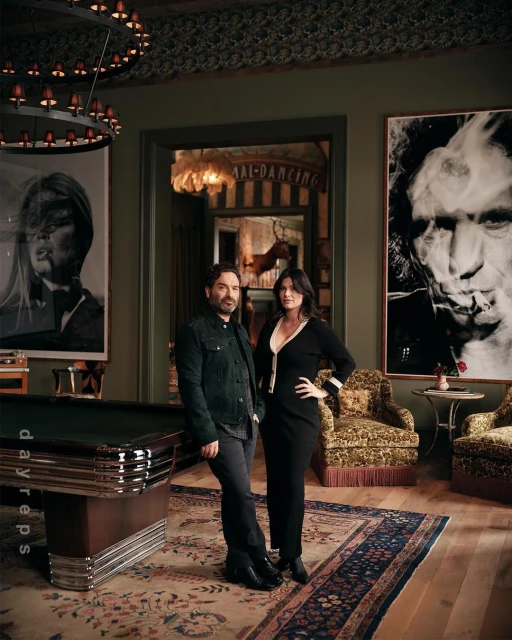 A man and a woman pose in an elegant interior with artwork, pool table, and ornate furnishings, captured with eye-level angle.