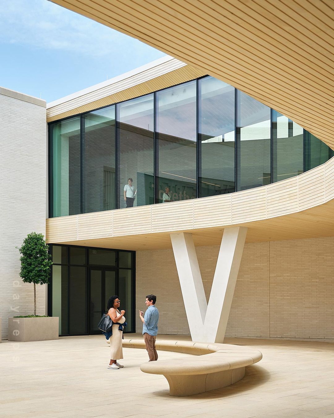 Two people stand in a modern building atrium with large windows, modern architecture, and neutral color scheme under a bright sky.