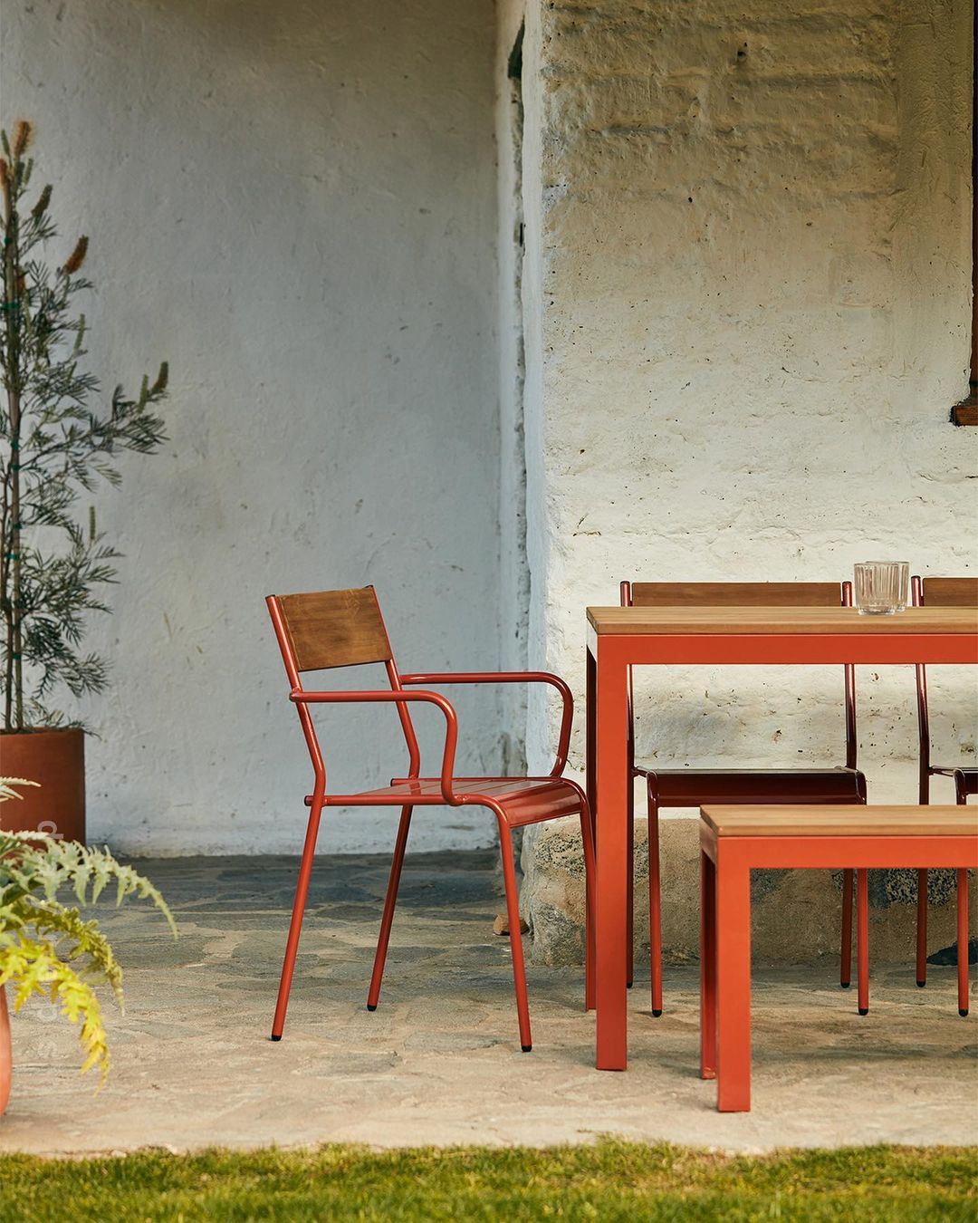Outdoor dining setup features a table, chairs, and bench with terracotta frames and wooden surfaces on a patio with greenery.