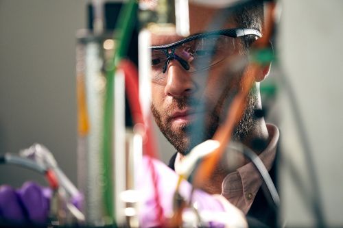 Close-up of an adult male wearing safety glasses, with assorted wires and equipment visible in the blurred foreground. 
