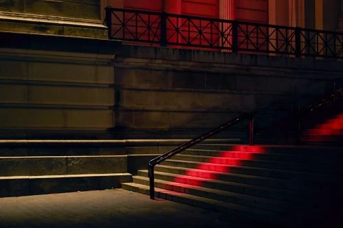 Exterior view of concrete stairs with metal railing illuminated by red light, set against a building facade at nighttime.