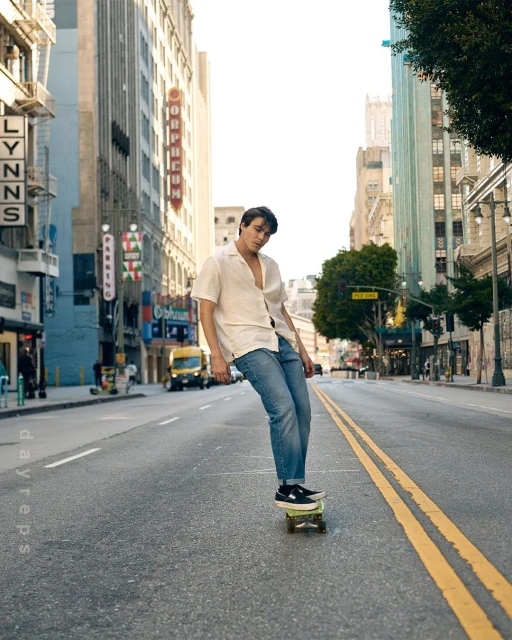 An adult male skateboarding on a city street with tall buildings, wearing a white shirt and blue jeans, during the daytime.