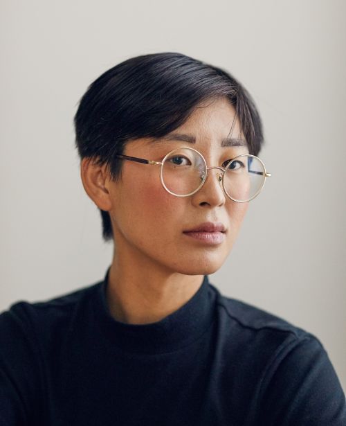 Portrait of an adult woman with short black hair and gold-rimmed glasses, wearing a black turtleneck shirt against a neutral background.
