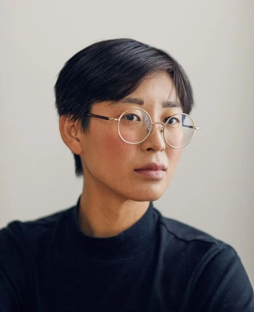 Portrait of an adult woman with short black hair and gold-rimmed glasses, wearing a black turtleneck shirt against a neutral background.