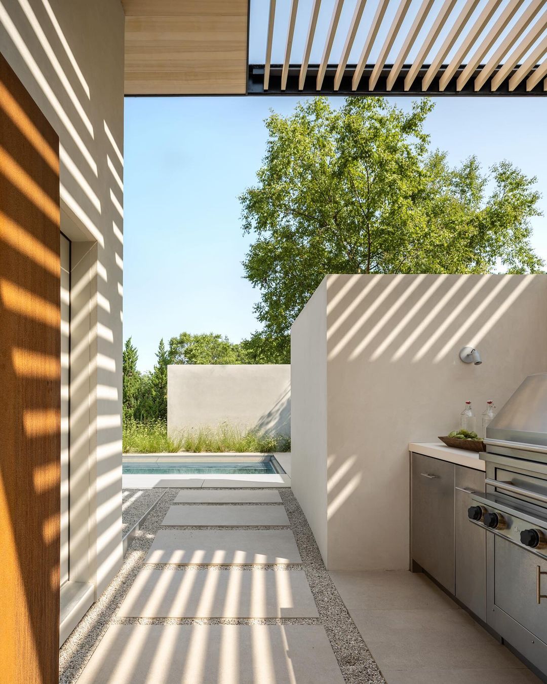 Modern outdoor kitchen and patio area with a stainless steel grill, white walls, and a pergola casting shadows on the stone floor.