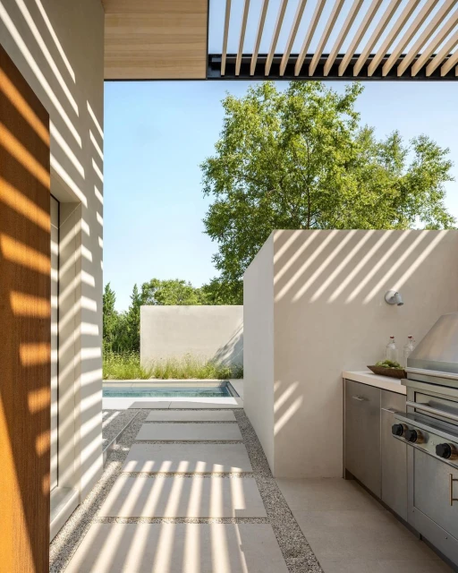 Modern outdoor kitchen and patio area with a stainless steel grill, white walls, and a pergola casting shadows on the stone floor.