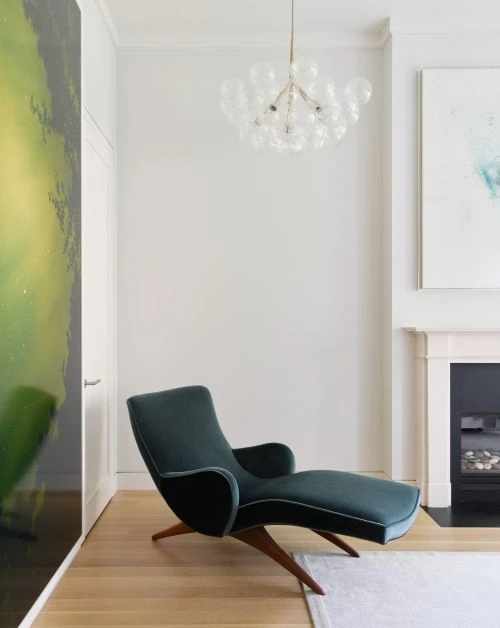 Modern living room featuring a teal chaise lounge with wood legs, a bubble chandelier, and abstract art on a light wood floor.
