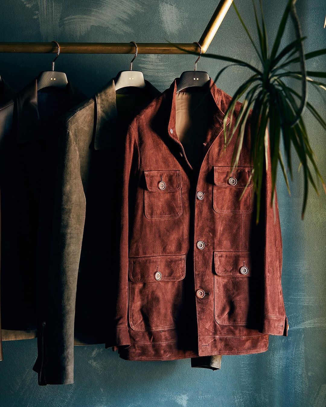 Hanging garments including a brown button-down jacket with pockets and other clothing on a metal rack with a houseplant.