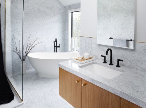 Modern bathroom features a white freestanding bathtub, marble walls, a wood vanity, and black fixtures in a well-lit interior space.