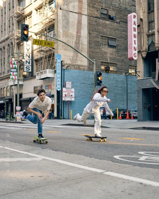 Two young adults, a man and a woman, are skateboarding on a city street, surrounded by buildings and traffic signals on a sunny day.