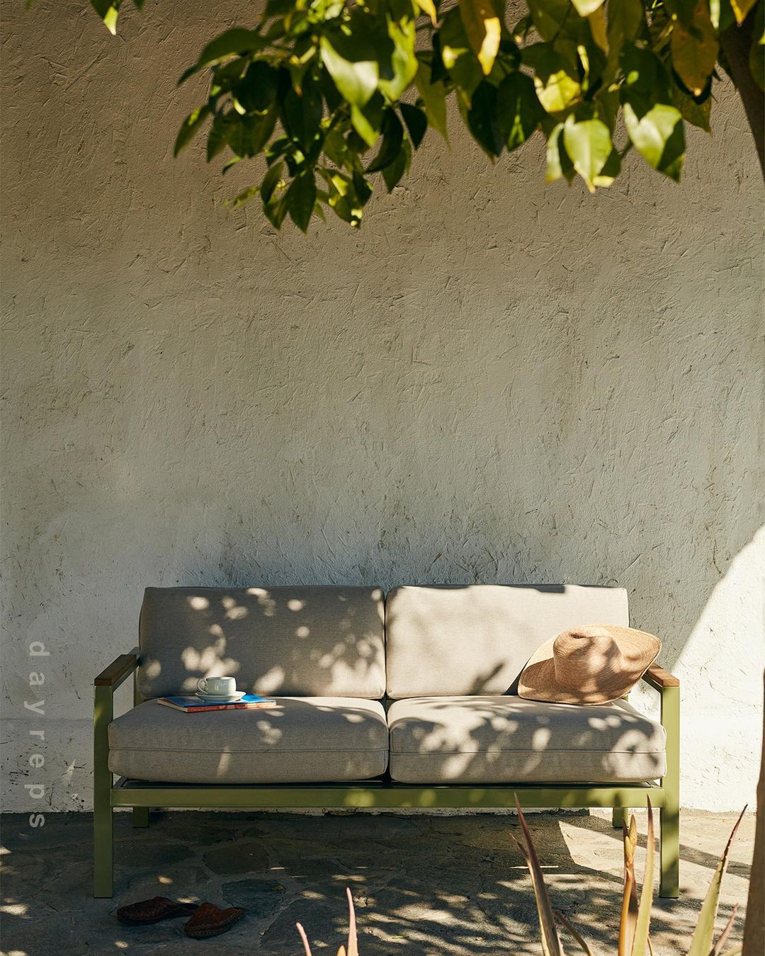 Outdoor seating area with a loveseat sofa, hat, drink, and reading materials set on a stone patio beneath a leafy tree canopy.