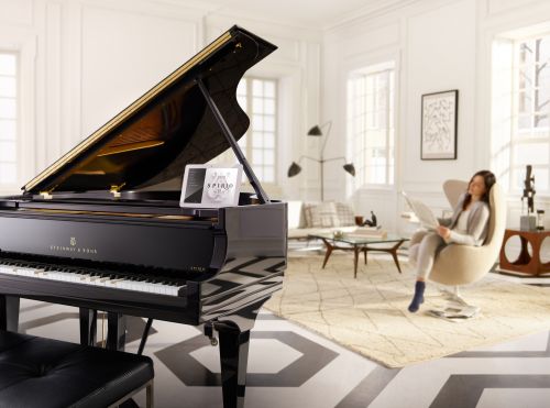 Grand piano with an open lid and a music book sits in a bright room with a woman reading in the background. Modern decor.