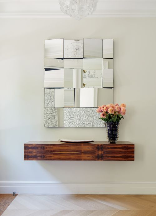 Interior view of a modern living room showcasing a mosaic mirror, floating wood shelf, a vase of pink flowers, and parquet flooring.