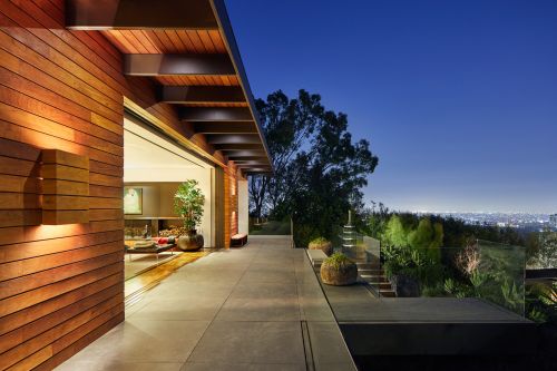 Exterior view of a modern house with wood siding, a patio with transparent guardrail, and a view of city lights at night.