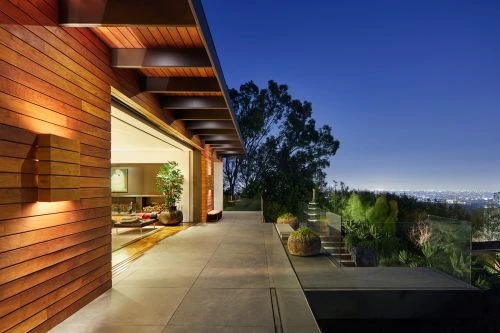 Exterior view of a modern house with wood siding, a patio with transparent guardrail, and a view of city lights at night.
