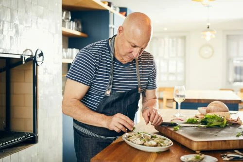 Adult male prepares oysters on a wooden counter in a well-lit kitchen wearing a striped shirt and a denim apron, indoor food preparation.