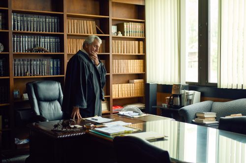 Man in robe standing in law office near desk and bookshelves filled with law books, next to windows, indoor setting.