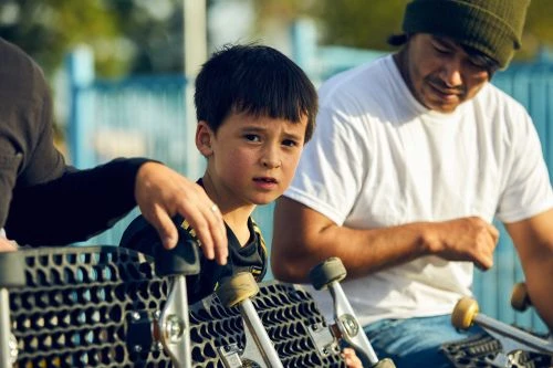 A young boy with a skateboard stands alongside an adult, wearing a beanie, and another person near a blue fence outdoors.
