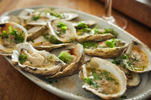 A platter of fresh, raw oysters on the half shell with green herbs, presented with natural light on a wooden table.