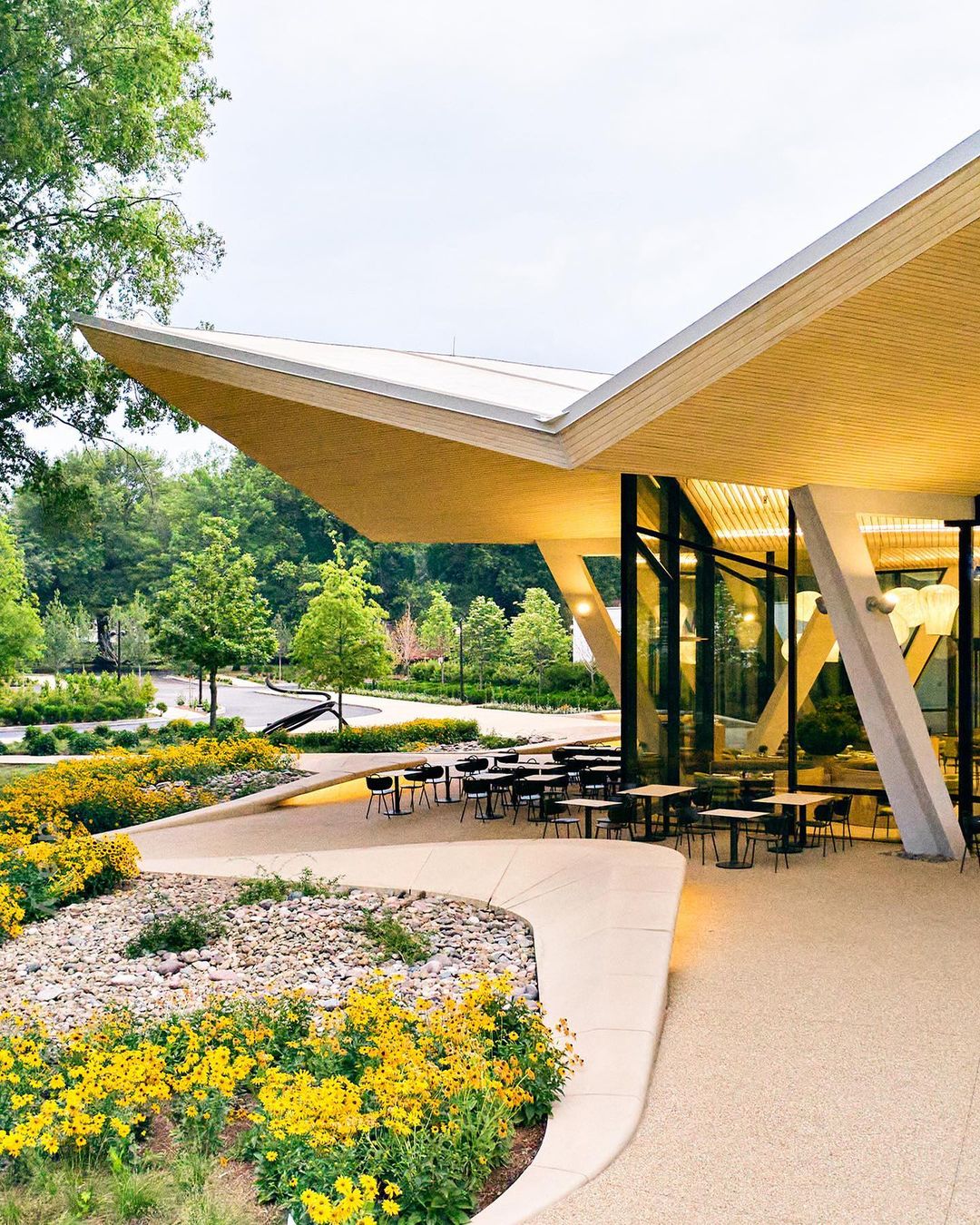 Exterior view of a modern building featuring large glass windows, a cantilevered roof, and a landscaped garden with yellow flowers and trees.