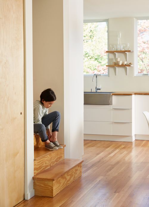 A child sitting on wooden stairs in a modern home tying their shoe laces, with a view to a contemporary kitchen and hardwood floors.