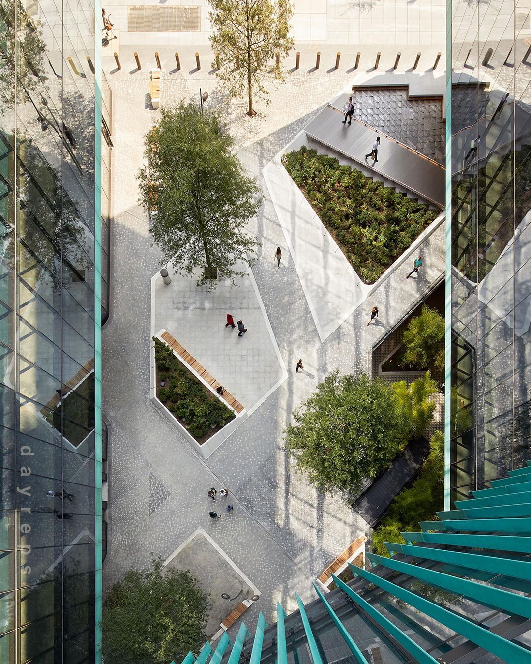 Aerial view of an outdoor public space with geometric landscaping, trees, benches, and pedestrians walking on paved paths between buildings.