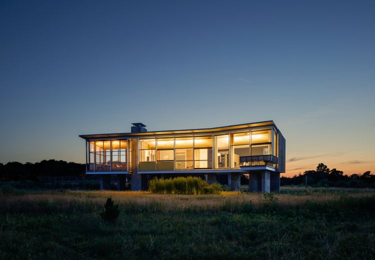 Exterior shot of modern home with large windows at dusk, set in a field with a darkening blue sky and warm interior lighting.