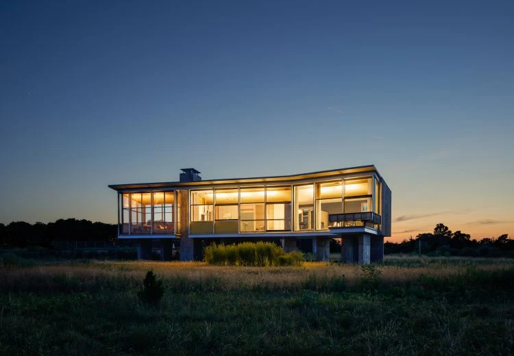 Exterior shot of modern home with large windows at dusk, set in a field with a darkening blue sky and warm interior lighting.