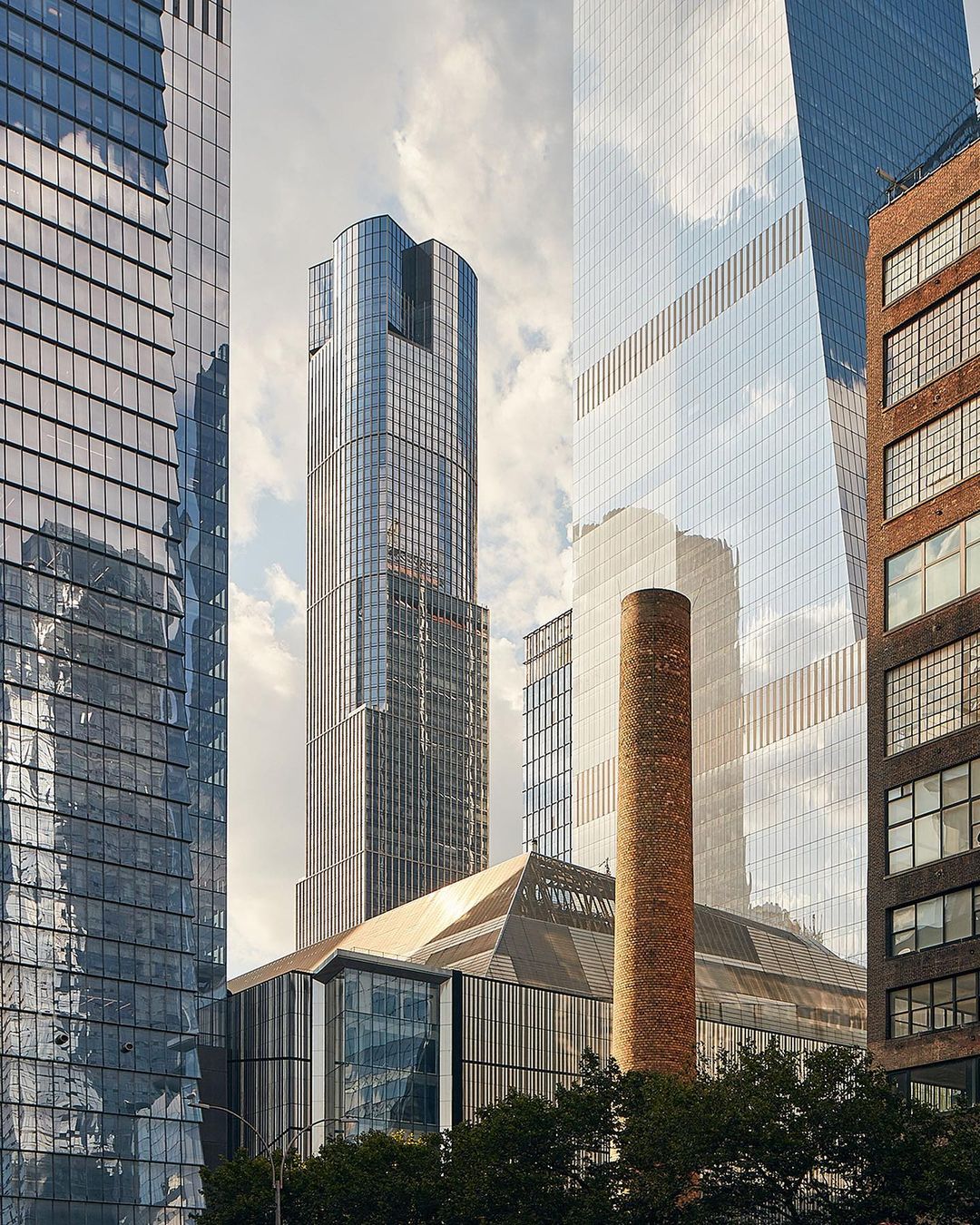 New York City architecture featuring glass skyscrapers, a brick smokestack, and lush green trees in an urban setting on a cloudy day.