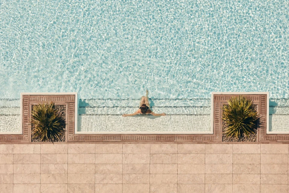 Overhead shot of a woman in a swimming pool with tile surround and planters with green spiky plants on either side of the steps.