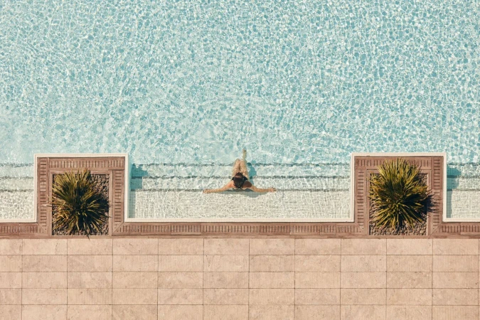 Overhead shot of a woman in a swimming pool with tile surround and planters with green spiky plants on either side of the steps.