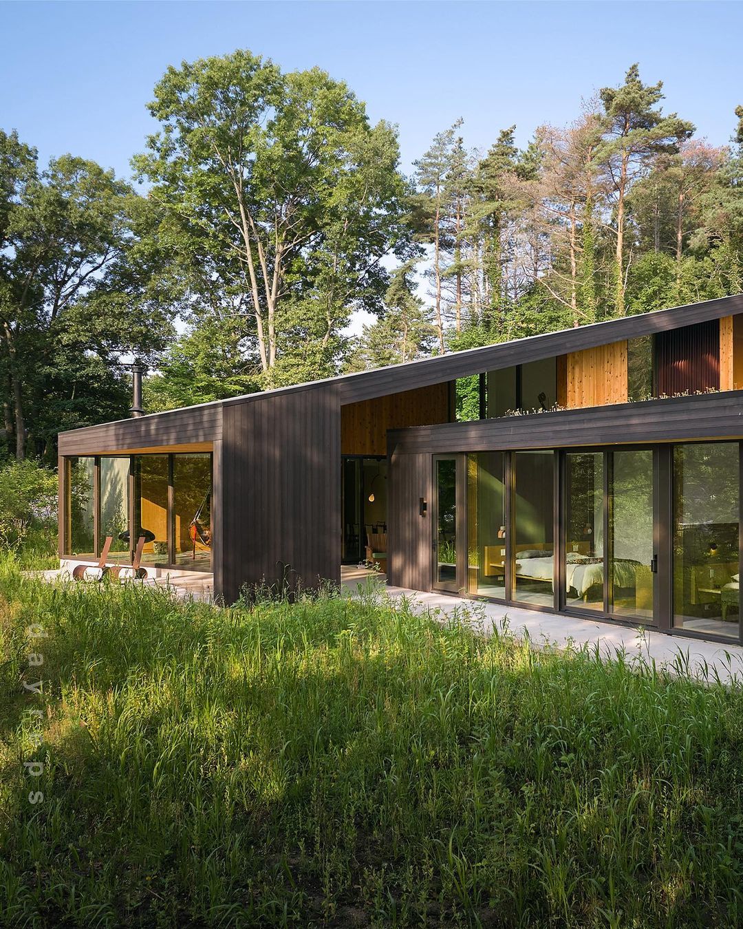 Modern architectural house with wood cladding and large glass windows nestled amongst green trees and meadow.