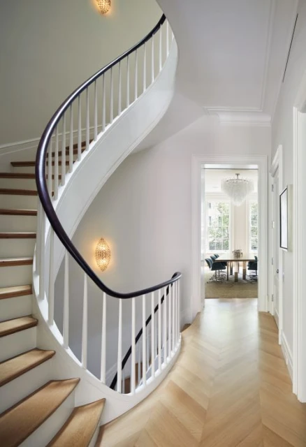 Interior view of a curved staircase with white railing and wooden steps, featuring herringbone wood flooring and soft, diffused lighting.