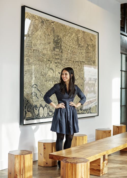 Woman in a blue dress standing in an interior space with a large framed drawing and wooden stools arranged around a table.