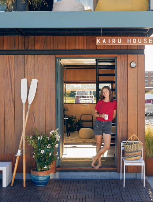 Adult woman standing in the doorway of a waterfront house, holding a mug with white paddles and a flower pot on the porch.