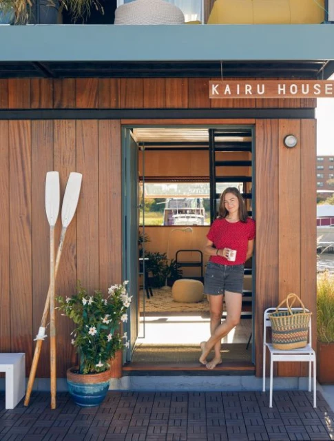 Adult woman standing in the doorway of a waterfront house, holding a mug with white paddles and a flower pot on the porch.