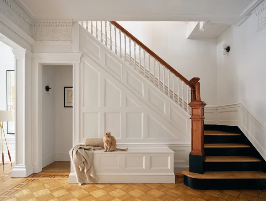 Feline sits on a cushioned bench in a bright hallway with wood floors, a staircase, and white molding details in a home interior.