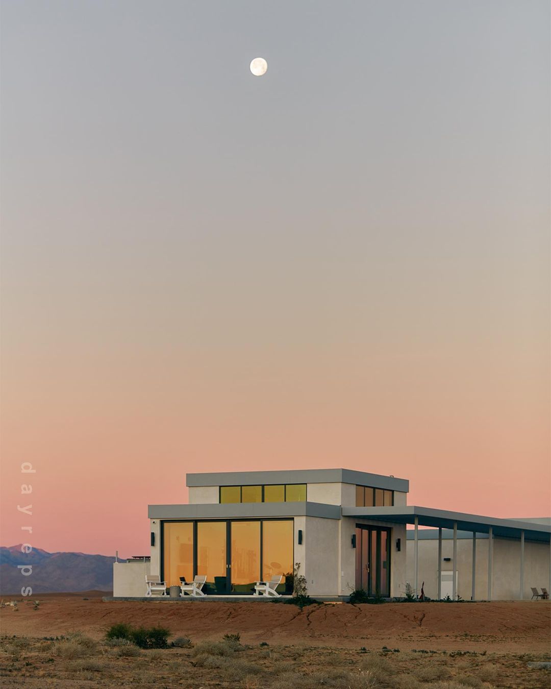 Modern architecture house with flat roof, reflected windows, outdoor furniture in desert landscape at dusk under a full moon.