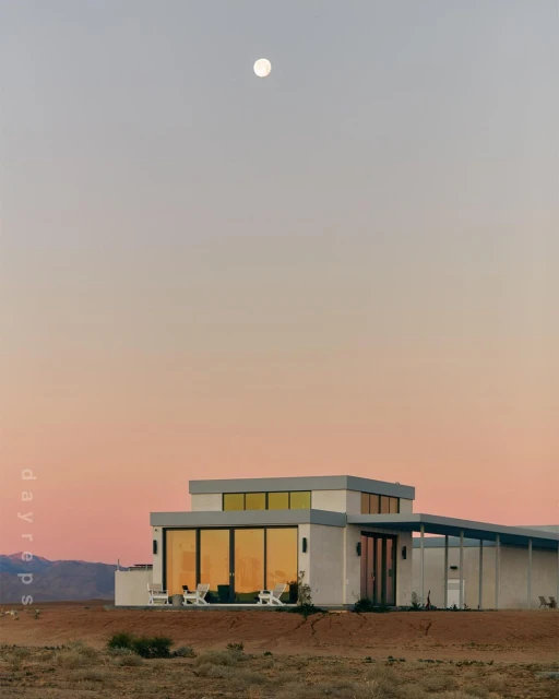 Modern architecture house with flat roof, reflected windows, outdoor furniture in desert landscape at dusk under a full moon.