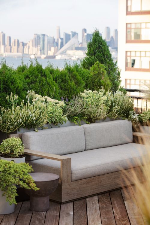 Outdoor sofa on wood deck surrounded by green plants and city view. Casual seating area on rooftop with grey cushions, exterior view.