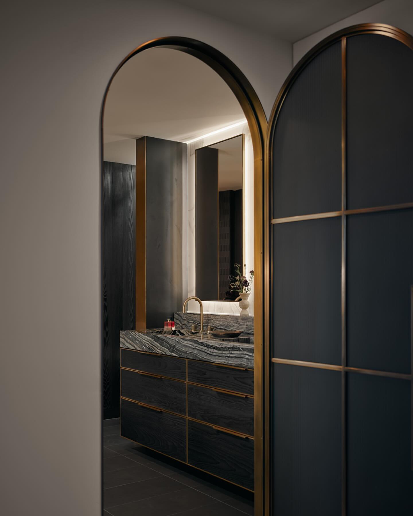Modern bathroom interior featuring a vanity with a stone countertop, black cabinets, and a gold faucet, viewed through arched doorways.